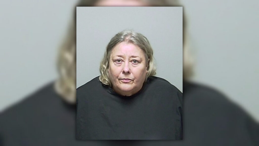 Booking photo of Christine Ann Woodworth, a woman with shoulder-length blond and gray hair wearing a dark top, against a gray background.